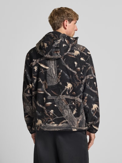 Männermodel trägt Fleecejacke mit Kapuze Modell 'Blevin' von Carhartt Work In Progress in braun-schwarzem Tarnmuster, Ganzkörperansicht Rückansicht. Gerader Schnitt, Zwei-Wege-Reißverschluss, Kapuze, Seitentaschen, Label-Patch, Rückenlänge bei Größe M 67 cm, Ärmellänge 71 cm.