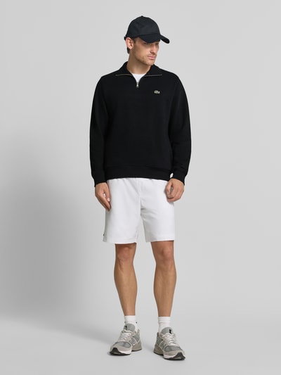 Männermodel trägt Regular Fit Troyer aus reiner Baumwolle von Lacoste in Schwarz, glatte Textur, kombiniert mit weißen Shorts, Ganzkörperansicht von vorne. Herren Troyer von Lacoste.