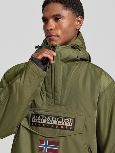 Männermodel trägt Jacke mit Kapuze und kurzem Reißverschluss von Napapijri in Olivgrün mit Pattentasche und Label-Patches, Nahaufnahme Oberkörper. Modell 'RAINFOREST NEXT', gerader Schnitt, Kapuze mit Tunnelzug und Kordelstoppern, sichtbarer Markenaufdruck.