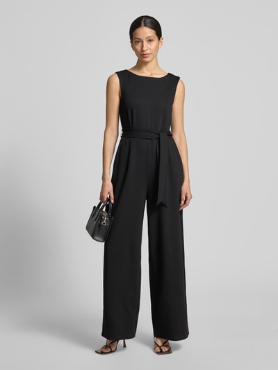 Vrouwenmodel draagt zwarte 'Jumpsuit aus Viskose-Mix mit Bindegürtel' van Jake*s Collection, viscosemix met stretch, regular fit, ronde hals, strikceintuur, ruglengte maat 36: 40 cm, borstwijdte maat 36: 30 cm, totale lengte maat 36: 150 cm, binnenbeenlengte maat 36: 80 cm, gladde lichte textuur, rechte lijnen, kleur effen zwart, full-body vooraanzicht.