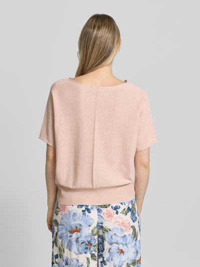 Modelka nosi sweter z dzianiny z dekoltem w łódkę i brustaszą 'Strickpullover mit U-Boot-Ausschnitt und Brusttasche' od Oui, bawełna, krój loose fit, rękaw o dł. 1/2, prążkowany dół, długość tyłu w rozmiarze 36: 52 cm, długość rękawów w rozmiarze 36: 31 cm, różowy, gładka tekstura, widoczne szerokie kwiatowe spodnie, zdjęcie całej sylwetki z tyłu.