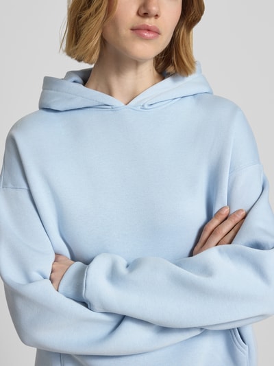 Modelka kobieca ma na sobie Regular Fit Hoodie mit Känguru-Tasche Modell 'Iconic' od Gina Tricot, bawełniana mieszanka, elastyczne wykończenia, kolor jasnoniebieski, z przodu kieszeń kangurka; zbliżenie na górną część ciała, widok z przodu.