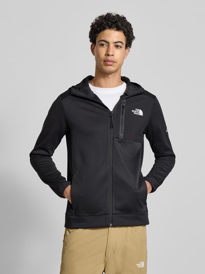 Männermodel trägt schwarze Sweatjacke mit Napoleontasche Modell 'MOUNTAIN ATHLETICS' von The North Face, Ganzkörperansicht von vorne. Passform: Körpernah, Kapuze, durchgehender Reißverschluss, Label-Details, elastische Abschlüsse, Rückenlänge bei Größe M 68 cm, Ärmellänge 67 cm.