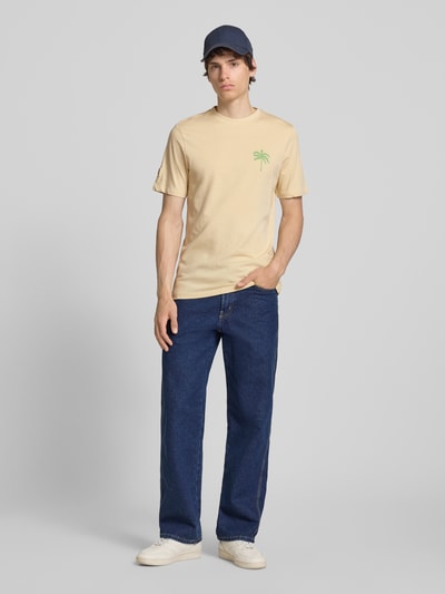 Männermodel trägt Herren T-Shirt mit Motiv-Print Modell 'SUN PALMS' von Blend, Reine Baumwolle, Regular-Fit, Rundhalsausschnitt, dezente Sandfarbe und grünes Print-Motiv, Rückenlänge bei Größe M: 73 cm, Ganzkörperansicht von vorne.