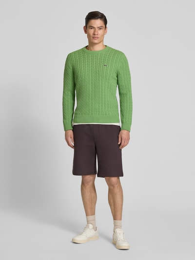 Männermodel trägt Regular Fit Strickpullover aus Baumwoll-Mix von Lacoste, Optik: Unifarben, Zopfmuster, Logo-Patch, Ganzkörperansicht von vorne.