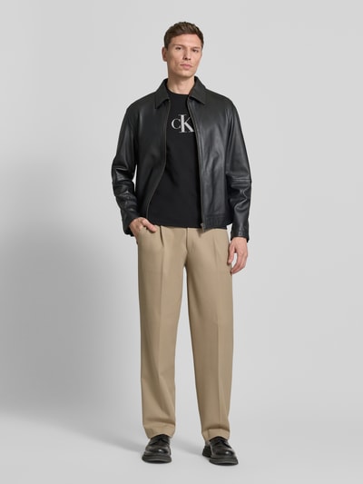 Männermodel trägt T-Shirt mit Rundhalsausschnitt und Logo von CK Calvin Klein in Schwarz, kombiniert mit beiger Hose, Ganzkörperansicht von vorne. Reine Baumwolle, Regular Fit, Logo-Detail, Rückenlänge bei Größe M: 70 cm.