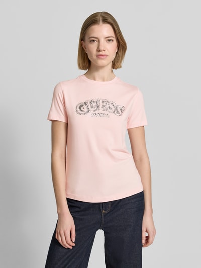Modelka nosi T-Shirt aus Viskose-Elasthan-Mix mit Logo od Guess Activewear, kolor jasnoróżowy, krój regular fit, okrągły dekolt, logo na przodzie, długość pleców w rozmiarze S: 53 cm, widok całej sylwetki z przodu.