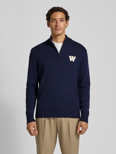 Männermodel trägt Strickpullover aus reiner Lammwolle mit Logo-Patch Modell 'Tay' von Wood Wood, navyblau, Regular Fit, Troyer-Kragen, Langarm, gerippte Abschlüsse, Ganzkörperansicht von vorne.