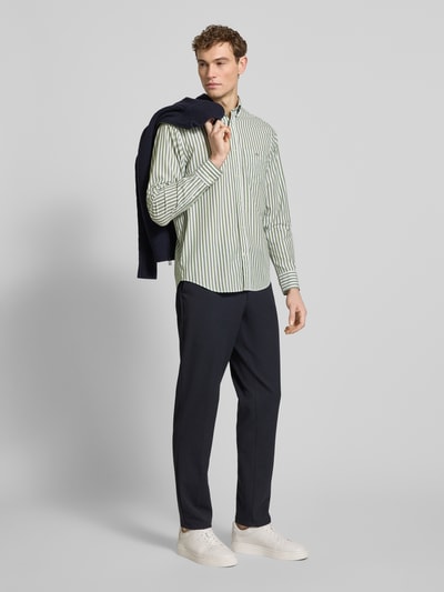 Męski model nosi Slim Fit Freizeithemd mit Brusttasche od Gant z czystej bawełny, pionowe zielone paski, krój slim fit, kieszeń na piersi, logo, długi rękaw, kołnierzyk button-down, zaokrąglony dół, długość pleców w rozmiarze M 77 cm, szerokość w rozmiarze M 46 cm, długość rękawa w rozmiarze M 65 cm, pełny widok z przodu.