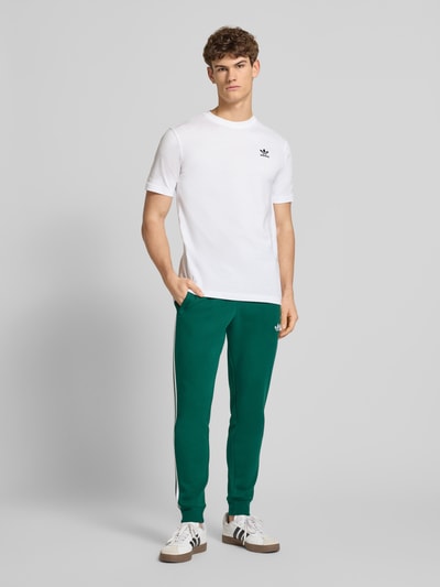 Männermodel trägt Sweatpants mit elastischem Bund von adidas Originals in Dunkelgrün, Baumwoll-Elasthan-Mix, lockerer Schnitt, Ganzkörperansicht von vorne.