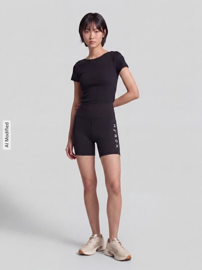 Frauenmodel trägt schwarze Elastische Sportshorts mit Logo-Print Modell 'PUMA X HYROX ESS SHORT' von PUMA, Damen Sportshorts, Ganzkörperansicht von vorne, KI-modifiziertes Bild.