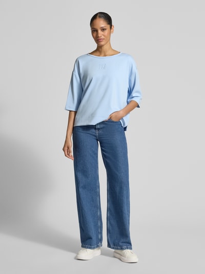 Frauenmodel trägt T-Shirt mit Rundhalsausschnitt und Logo von monari in Hellblau, Relaxed Fit, glatte Textur, Ganzkörperansicht von vorne.