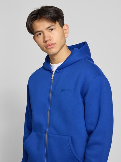Männermodel trägt Sweatjacke mit Känguru-Tasche von REVIEW in Blau aus Baumwoll-Mix, Relaxed-Fit, Reißverschluss, Nahaufnahme Oberkörper.