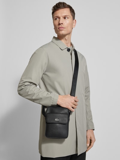 Männermodel trägt Umhängetasche mit Label-Applikation von Lacoste aus schwarzem, genarbtem Leder, rechteckige Form, vorne, Nahaufnahme Oberkörper.