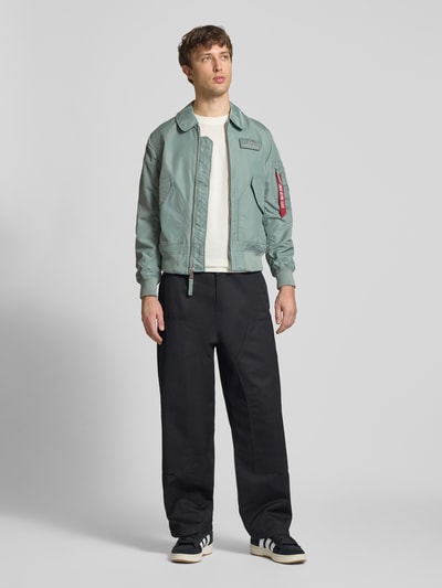 Męski model nosi Regular Fit Bomberjacke mit Logo-Patch od Alpha Industries, kolor zielony, wykładany kołnierz, nakładane kieszenie, zamek błyskawiczny, elastyczne wykończenia, długość tyłu w rozmiarze M 66 cm, szerokość ramion w rozmiarze M 44 cm, szerokość klatki piersiowej w rozmiarze M 56 cm, długość rękawów w rozmiarze M 66 cm, ujęcie całej sylwetki z przodu.