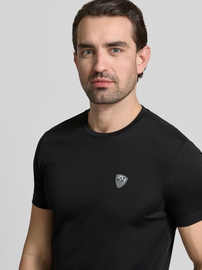Mężczyznamodel nosi T-Shirt aus Viskose-Mix mit Label-Patch von EA7 Emporio Armani, czarny, krój slim fit, okrągły dekolt, zbliżenie na górną część ciała