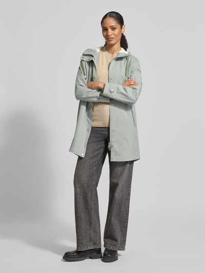Frauenmodel trägt Jacke mit Ärmelriegeln von NAUTGÅRD in hellgrün mit Kapuze, Knopfleiste und Reißverschlusstaschen, Ganzkörperansicht von vorne.