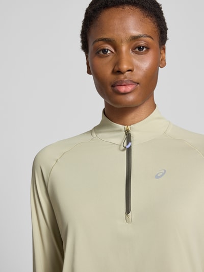 Frauenmodel trägt Longsleeve mit Troyer-Kragen Modell 'NAGINO' von ASICS TIGER in hellem Beige, reflektierender Logo-Print, Nahaufnahme Oberkörper.