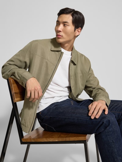 Männermodel trägt Regular Fit Jacke aus Lyocell-Mix von Guess in Olivgrün, Umlegekragen, Reißverschlusstaschen, elastischer Bund, Nahaufnahme Oberkörper.