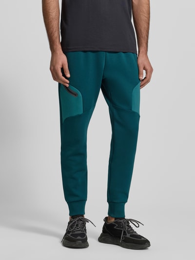 Männermodel trägt Regular Fit Sweatpants mit Logo-Print Modell 'Unstoppable' von Under Armour, Elastischer-Material-Mix, Beintasche mit Reißverschluss, elastischer Bund und Beinabschlüsse, Innenbeinlänge 73 cm, Farben Blau und Dunkelgrün, Ganzkörperansicht von vorne.