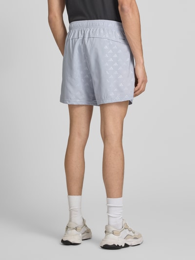 Mężczyzna nosi Regular Fit szorty z wzorem z logo na całej powierzchni model 'WE BRLO SHO' od adidas Training, efekt: jednolity kolor, krój regular fit, detal z logo, elastyczny ściągacz, boczne kieszenie zapinane na zamek błyskawiczny, wykończenie materiału: satyna, całkowity widok z tyłu.