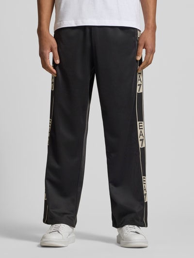 Männermodel trägt Trainingshose mit seitlichen Knopfleisten von EA7 Emporio Armani in Schwarz, Relaxed-Fit, Ganzkörperansicht von vorne.