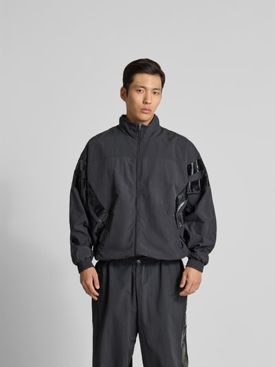 Männermodel trägt Trainingsjacke mit Stehkragen von VETEMENTS in Dunkelgrau mit glänzenden Akzenten, lockerer Schnitt, Ganzkörperansicht von vorne, Relaxed Fit, Zweiwege-Reißverschluss, elastische Abschlüsse, Eingrifftaschen, Rückenlänge 74 cm, Schulterbreite 50 cm, Ärmellänge 73 cm.