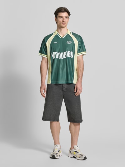 Männermodel trägt Poloshirt mit Label-Print Modell 'Taka' von WOODBIRD, lockere Passform, kurzärmelig, Polokragen, Ganzkörperansicht von vorne.