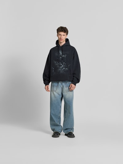 Männermodel trägt Oversized Hoodie aus Baumwoll-Mix von VETEMENTS in Schwarz mit Destroyed-Look, Motiv-Print und Känguru-Tasche, Ganzkörperansicht von vorne