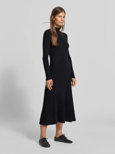 Frauenmodel trägt Strickkleid aus Viskose-Mix in Midilänge von MYON, schwarze Farbe, eng anliegend, A-Linie, Stehkragen, Ripp-Optik, Langarm, Ganzkörperansicht von vorne.
