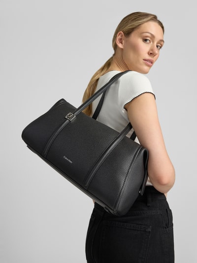 Vrouwenmodel draagt zwarte Henkeltasche met labeldetail van CK Calvin Klein voor dames, grof korrelige textuur, geheellichaamsaanzicht achterkant