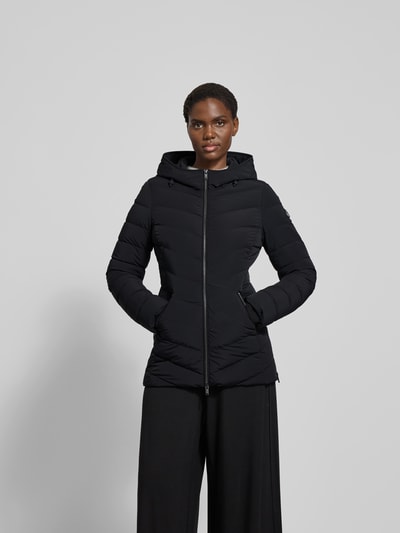 Frauenmodel trägt Daunenjacke mit Kapuze von Moose Knuckles in Schwarz, Material-Mix mit Stretch-Anteil, taillierter Schnitt, Reißverschluss, Reißverschlusstaschen, Rückenlänge 75 cm, Brustweite 48 cm, Ärmellänge 65 cm, Ganzkörperansicht von vorne.