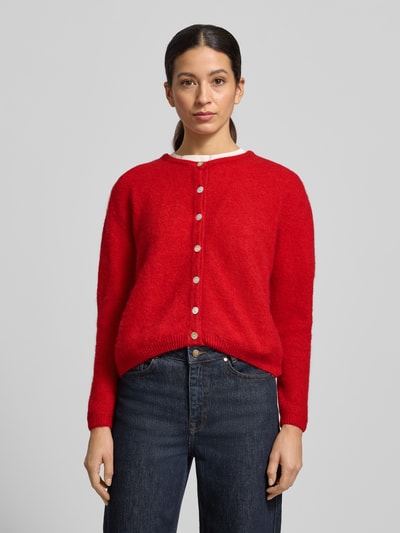 Frauenmodel trägt Strickjacke mit Knopfleiste Modell 'VITOW' von American Vintage in Rot mit gerippten Abschlüssen, Rundhalsausschnitt, lockerer Schnitt, Alpaka-Lanawoll-Mix mit Stretch, Jeans, Ganzkörperansicht von vorne.