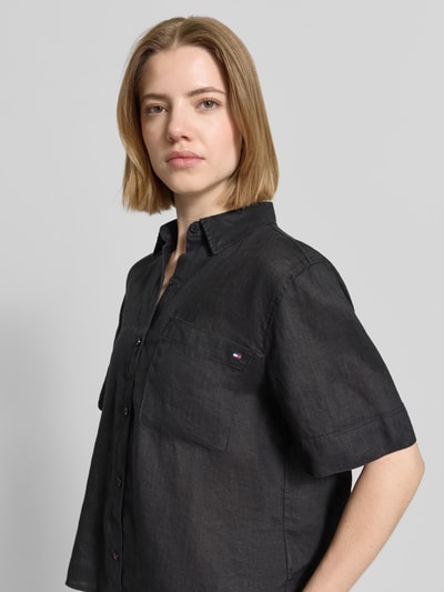 Frauenmodel trägt Regular Fit Leinenbluse mit Logo-Stitching von Tommy Hilfiger in Schwarz, kurzer Ärmel, Hemdblusenkragen, reine Leinenstruktur, Brusttasche, Nahaufnahme Oberkörper.