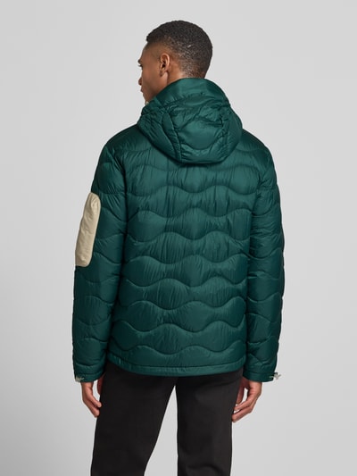 Männermodel trägt grüne Daunenjacke mit Kapuze von PEAK PERFORMANCE, beige Details, gerader Schnitt, Ganzkörperansicht Rückansicht.