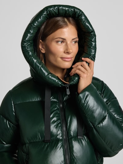 Frauenmodel trägt Steppmantel mit Kapuze und Label-Patch Modell 'ISABEL' von Save The Duck in dunklem Grün mit glatter, gesteppt glänzender Textur, Nahaufnahme Oberkörper. Rückenlänge 90 cm, Schulterbreite 45 cm, Brustweite 54 cm, Ärmellänge 64 cm, gerader Schnitt, Zweiwege-Reißverschluss, seitliche Reißverschlusstaschen.