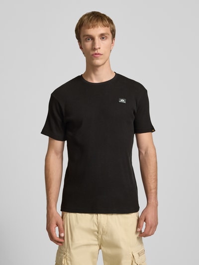 Männermodel trägt T-Shirt mit Label-Patch von Alpha Industries in Schwarz, Reine Baumwolle, Körpernahe Passform, Rundhalsausschnitt, Ganzkörperansicht von vorne.