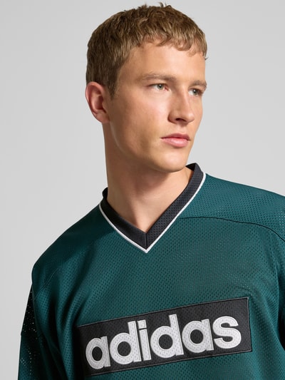 Männermodel trägt Longsleeve mit Label-Detail von adidas Originals, dunkelgrün, V-Ausschnitt, Lochstickerei, Nahaufnahme Oberkörper.