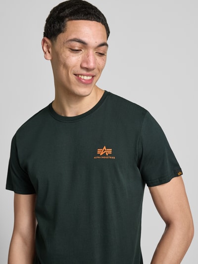 Mężczyznamodel nosi t-shirt z bawełny w kolorze ciemnozielonym z detalami z logo, model 'BASIC' od Alpha Industries, okrągły dekolt, prosty krój, długość tyłu w rozmiarze M: 67 cm, zbliżenie na górną część tułowia.