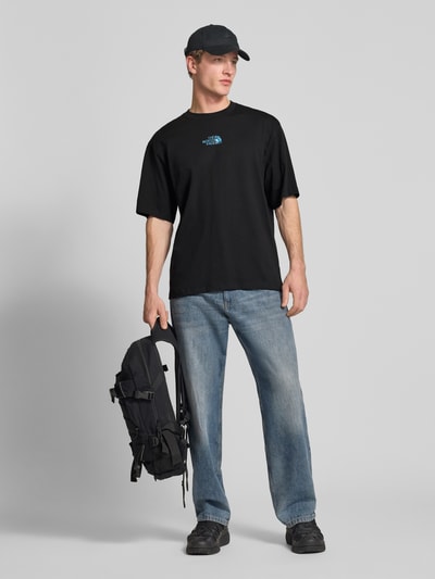 Męski model nosi Oversized T-Shirt mit Logo und Rundhalsausschnitt von The North Face, czarny kolor, czysta bawełna, fason oversized, logo na piersi, okrągły dekolt, długość tyłu w rozmiarze M: 73 cm, widok całej sylwetki z przodu.