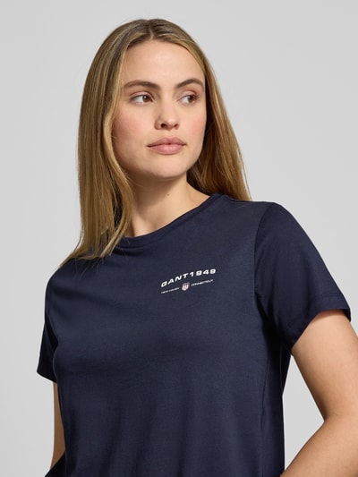Modelka kobieca ma na sobie granatowy T-Shirt z nadrukiem Gant, z bawełny, regular fit, okrągły dekolt, zbliżenie górnej części ciała.