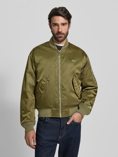 Männermodel draagt Regular Fit Bomberjack uit satijn van Lacoste in effen olijfgroen met logopatch, zijpaspelzakken, waterafstotend, warmte-isolatie, collegekraag, ritssluiting, geribde boorden, Ganzkörperansicht van voren.