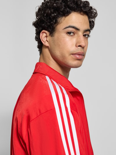 Męski model nosi Sweatjacke mit Stehkragen od adidas Originals, materiał z domieszką stretchu, czerwony z białymi paskami, widok z przodu, zbliżenie na górę ciała.