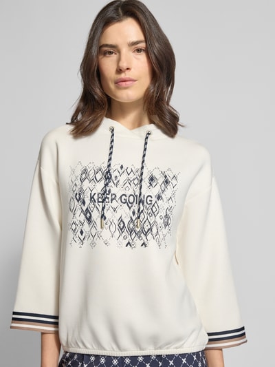 Modelka nosi Bluza 'Sweatshirt aus Viskose-Mix mit 3/4-Arm' od Betty Barclay, mieszanka wiskozy z dodatkiem streczu, krój relaxed fit, nadruk z napisem, kaptur z tunelem, rękaw o dł. 3/4, kremowe i granatowe odcienie, tekstura gładka, widok na górną część ciała z przodu.