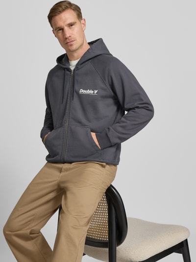 Męski model nosi Sweatjacke mit Kapuze und Reißverschluss Modell 'Sven' od Wood Wood, bawełna, relaxed fit, nadruk, kieszeń, widok całej sylwetki z przodu.
