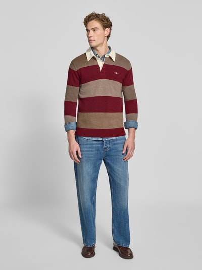Männermodel trägt Regular Fit Pullover aus reiner Baumwolle von GANT in Rot und Braun mit Streifen, blaue Jeans, Ganzkörperansicht von vorne.