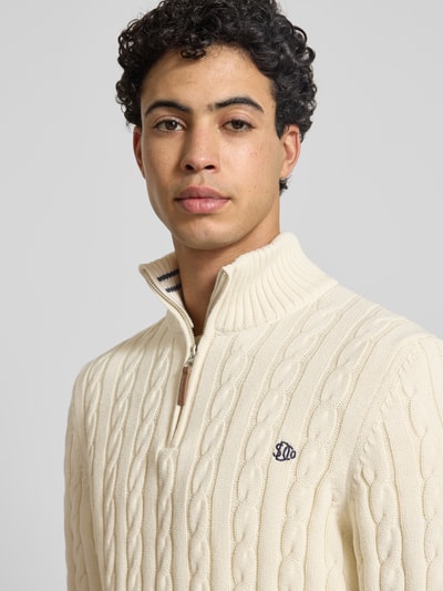 Männermodel trägt Strickpullover aus reiner Baumwolle von Superdry in Creme mit Zopfmuster, Troyer-Kragen und Label-Detail, Nahaufnahme Oberkörper.
