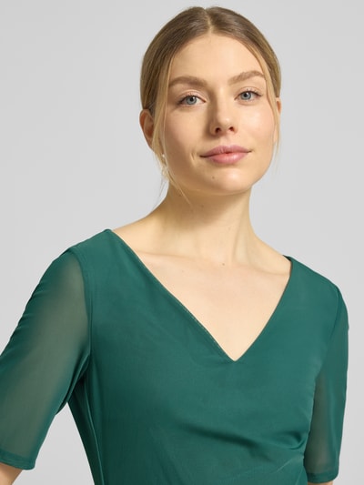 Frauenmodel trägt Regular Fit Midikleid in semitransparentem Design von Vera Mont in Grün, V-Ausschnitt, Kurzarm, Nahaufnahme Oberkörper. Elastischer Material-Mix, Nahtreißverschluss, blickdichter Unterrock, Rückenlänge 94 cm, Brustweite 37 cm, Ärmellänge 28 cm.