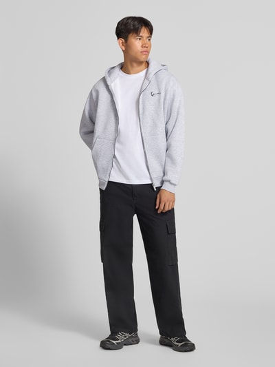 Männermodel trägt Sweatjacke mit Label-Stitching und Känguru-Tasche von Karl Kani, grau, Baumwoll-Mix, Reißverschluss, Ganzkörperansicht von vorne.