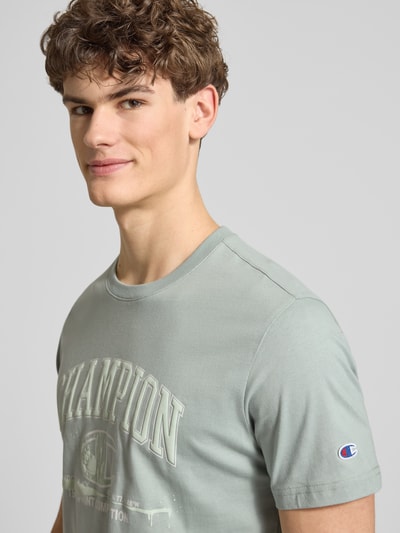 Mężczyzna nosi T-Shirt mit Label-Print und Rundhalsausschnitt von CHAMPION, czysta bawełna, krój regular fit, nadruk z logo, okrągły dekolt, długość pleców w rozmiarze M: 69 cm, zbliżenie na górną część ciała z przodu.